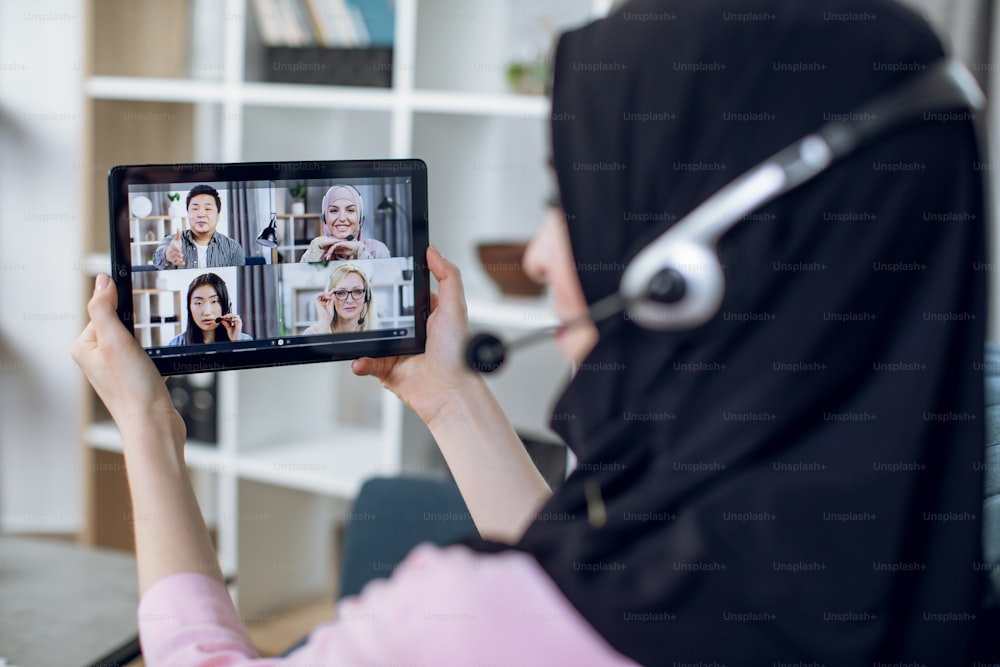 Young woman in hijab and headset having business meeting with international partners through video call. Female freelancer using digital tablet for online conversation with colleagues.