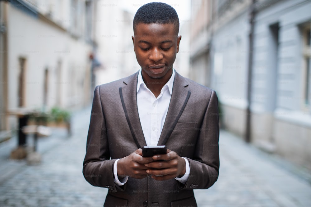 Confident afro american businessman in stylish suit standing on city street and using smartphone. Handsome man solving working issues online with help of modern gadget.