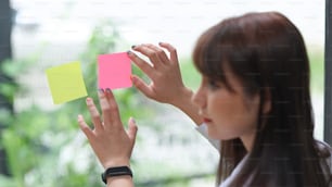 Primer plano de una empleada que pone notas adhesivas de colores en una pared de vidrio.