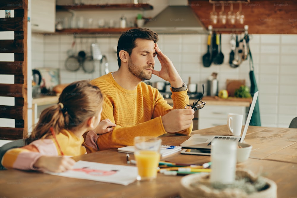 Male entrepreneur working on laptop and holding his head in pain while being with his daughter at home.