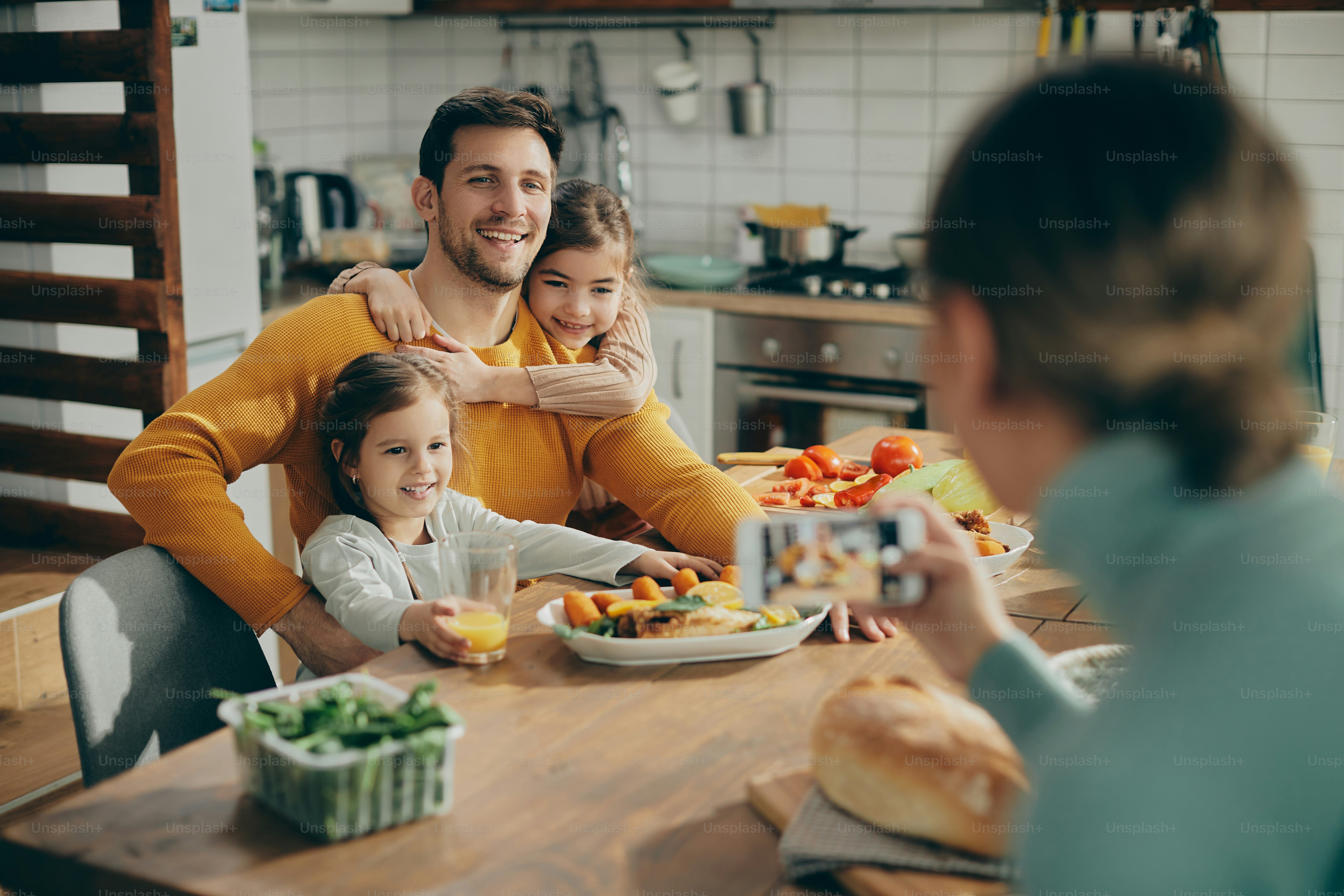 Happy father and daughters embracing while mother photographing them during a meal at dining table.
