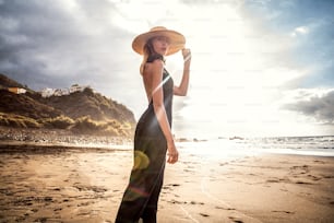 Modische Frau bei einem Sonnenuntergangsspaziergang am Strand. Sommerliche Inselatmosphäre. Weibliches Model mit elegantem Hut und schwarzem Kleid.