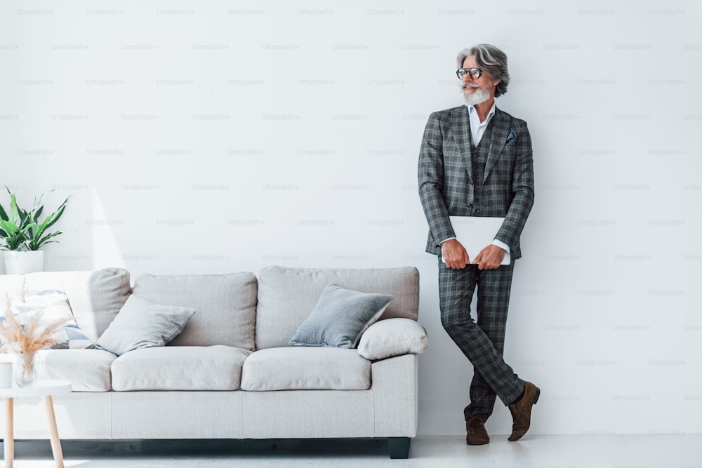 Senior stylish modern man with grey hair and beard indoors standing near wall with laptop in hands.