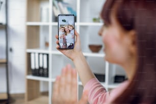 Young woman holding smartphone while talking with sister and her two cute kids. Pretty female having video call with relatives while staying at home.