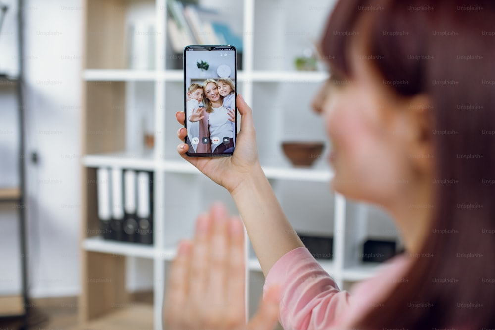 Young woman holding smartphone while talking with sister and her two cute kids. Pretty female having video call with relatives while staying at home.