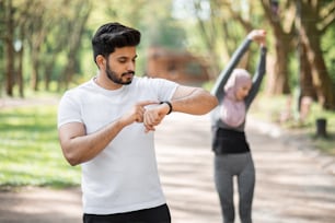 Hübscher muslimischer Mann, der App für das Training auf der Smartwatch verwendet, während die Frau im Hijab den Körper auf dem Hintergrund streckt. Junges aktives Paar, das im Freien trainiert.