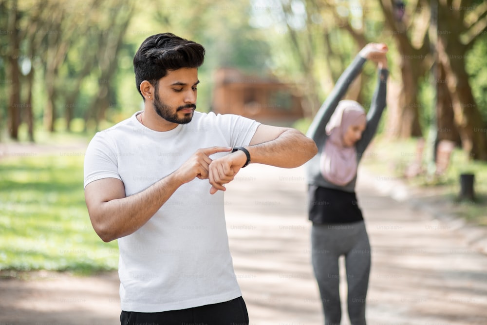 Handsome muslim man using app for training on smart watch while woman in hijab stretching body on background. Young active couple enjoying outdoors workout.