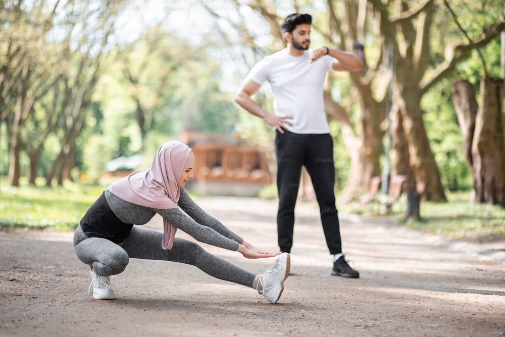 Mujer joven activa con hiyab haciendo ejercicios flexibles mientras un guapo chico árabe parado detrás y mirando un reloj inteligente. Concepto de personas, tecnología y actividad.