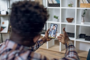 若い黒人男性は家に座って、アジアの雇用主とのオンライン就職面接に最新のスマートフォンを使用しています。リモートワーク、人、テクノロジーのコンセプト。