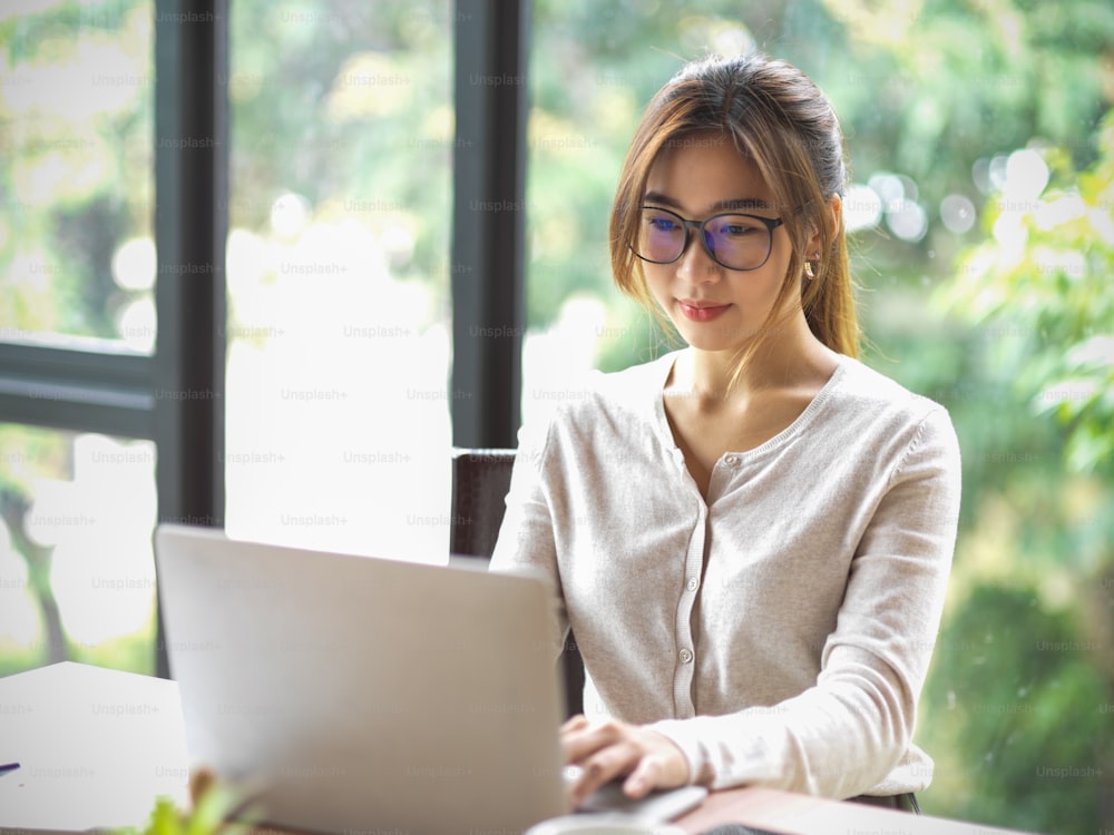 Portrait of beautiful asian businesswoman with eyeglasses working on laptop at office with blurred natural in the background