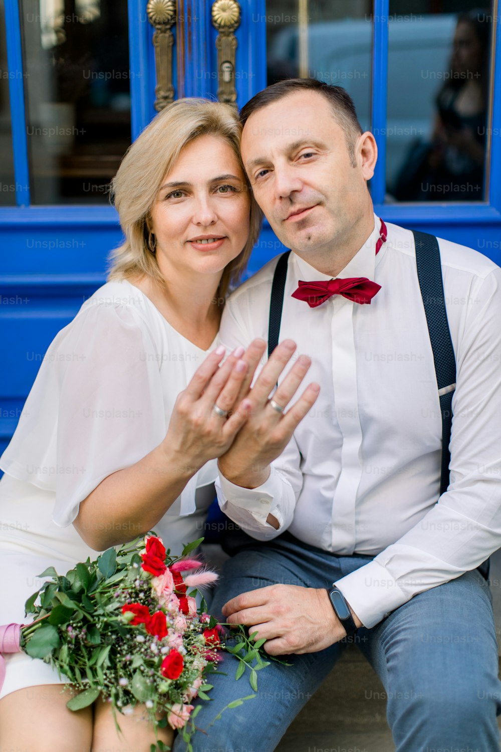 Love through the years. Happy smiling mature couple, sitting in front of blue vintage doors in the city street, showing their hands with the wedding rings. Focus on the faces