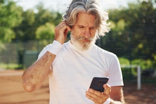 Mit Telefon und Kopfhörer. Senior modern, stilvoller Mann tagsüber im Freien auf dem Sportplatz.