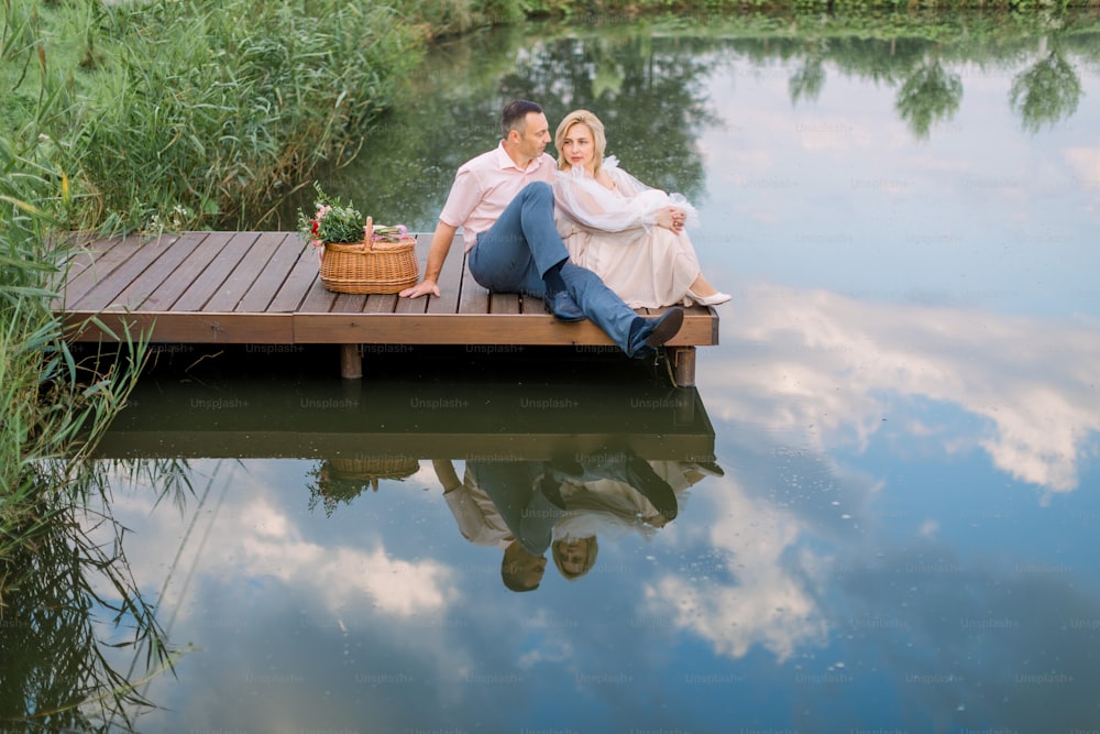 Happy romantic mature couple sitting on a wooden bridge near lake or pond outdoors, hugging and enjoying their date. Wedding anniversary celebration