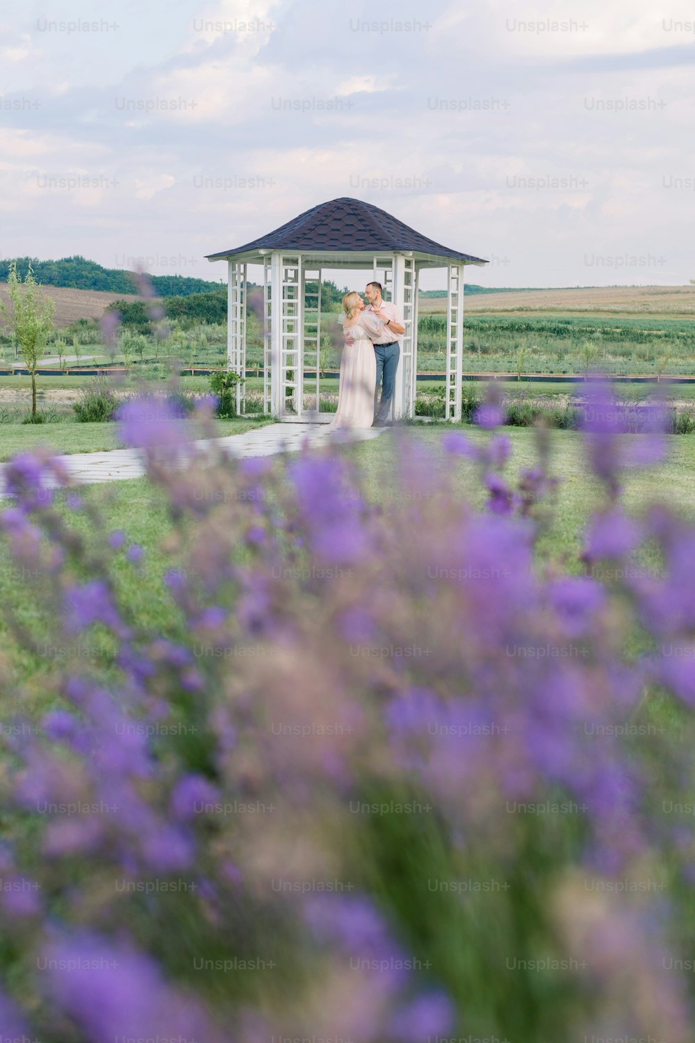 View through the flowering lavender of happy mature couple, man and woman in elegant clothes, standing near wooden gazebo on the shore of beautiful lake. Romantic date on the shore of the pond.