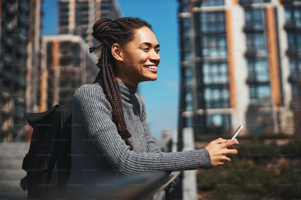 Portrait d’une touriste souriante et satisfaite avec un smartphone dans les mains, les coudes appuyés sur la rampe