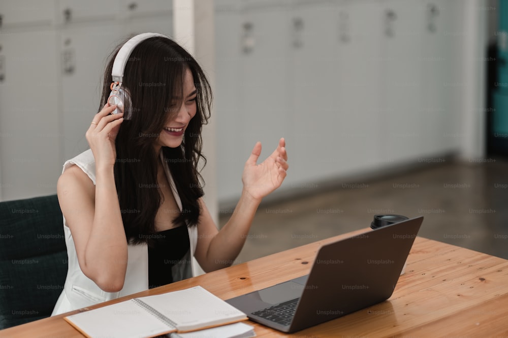Smiling girl student wear wireless headphone study online with teacher, happy young asian woman learn language listen lecture watch webinar write notes look at laptop sit in cafe, distant education
