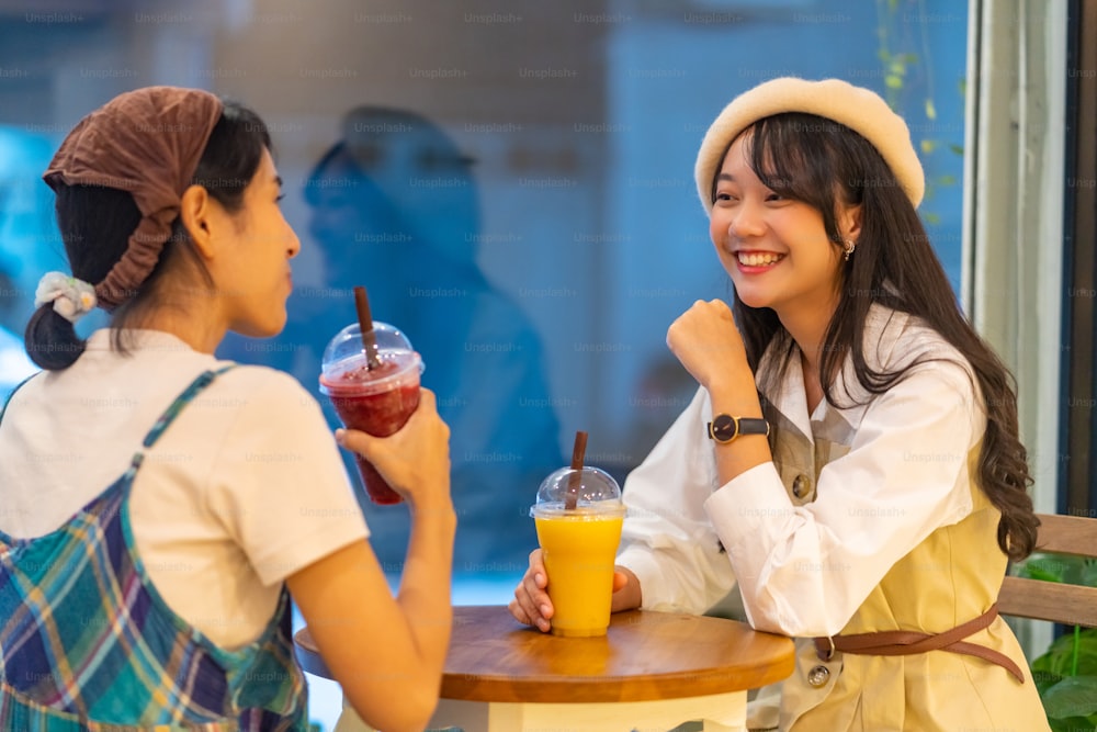 Young beautiful Asian woman friends sitting by the window in cafe with drinking fruit juice and talking together with happiness. Attractive female friendship enjoy leisure activity and meeting in the city