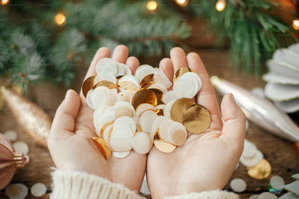 Manos sosteniendo confeti dorado sobre fondo de adornos navideños, adornos y ramas de pino en luces sobre madera rústica. Espacio para el texto. Felices Fiestas. Saludos de temporada