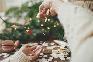 Hands tossing golden confetti on background of christmas lights, decorations, baubles and pine branches on rustic wood. Space for text. Happy holidays. New year and xmas party. Festive mood