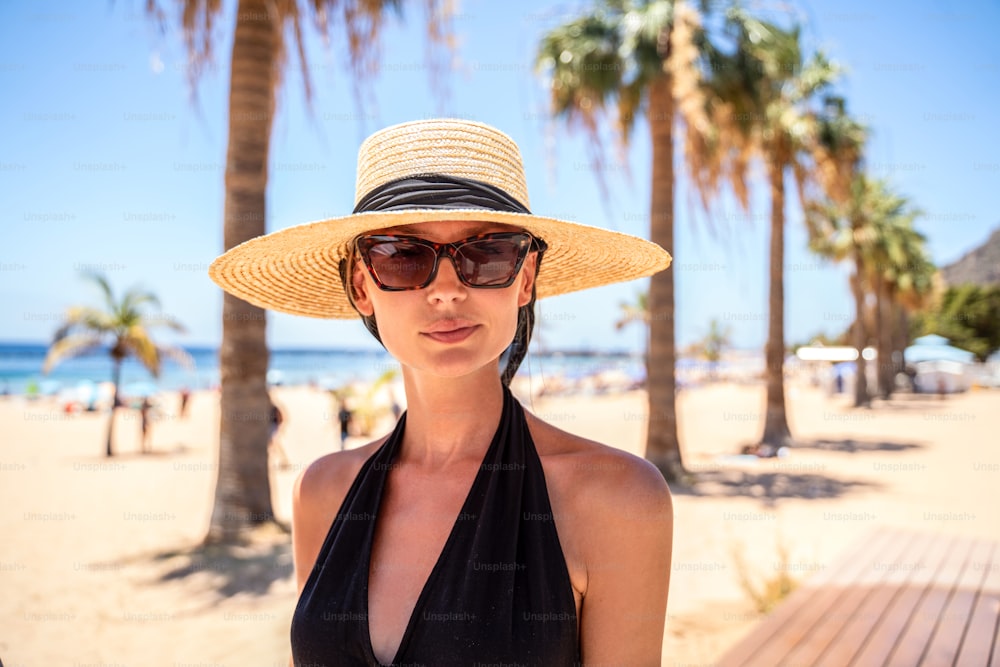 Foto de verão de mulher feliz usando chapéu e óculos escuros andando na praia. Palmas ao fundo.