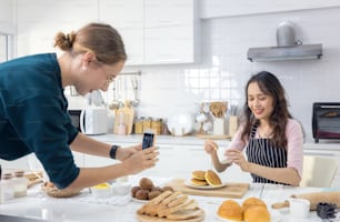 Beautiful woman pastry chef kneading bread dough on the worktop, as another female snaps a snapshot of her doing so.