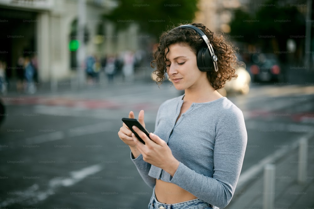 A young woman with headphones on, waiting on a crosswalk and choosing music on the phone.