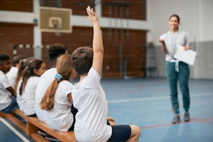 Vue arrière d’un élève du primaire levant le bras pour répondre à la question de l’enseignant pendant qu’il suivait un cours d’éducation physique au gymnase de l’école.