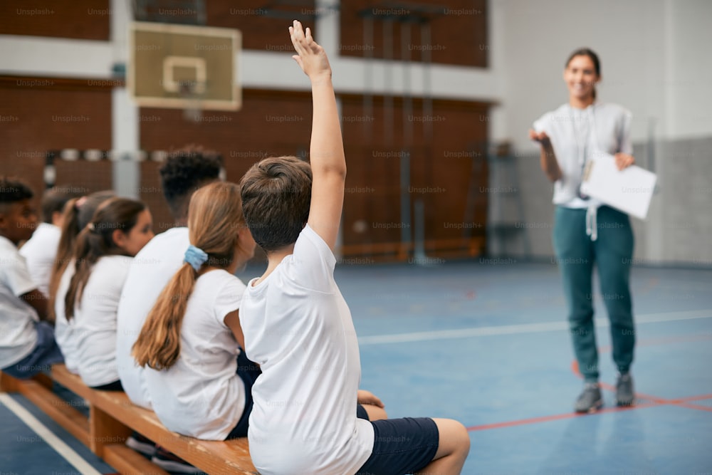 Vista posterior de un estudiante de primaria levantando el brazo para responder la pregunta del maestro mientras tiene clase de educación física en el gimnasio de la escuela.