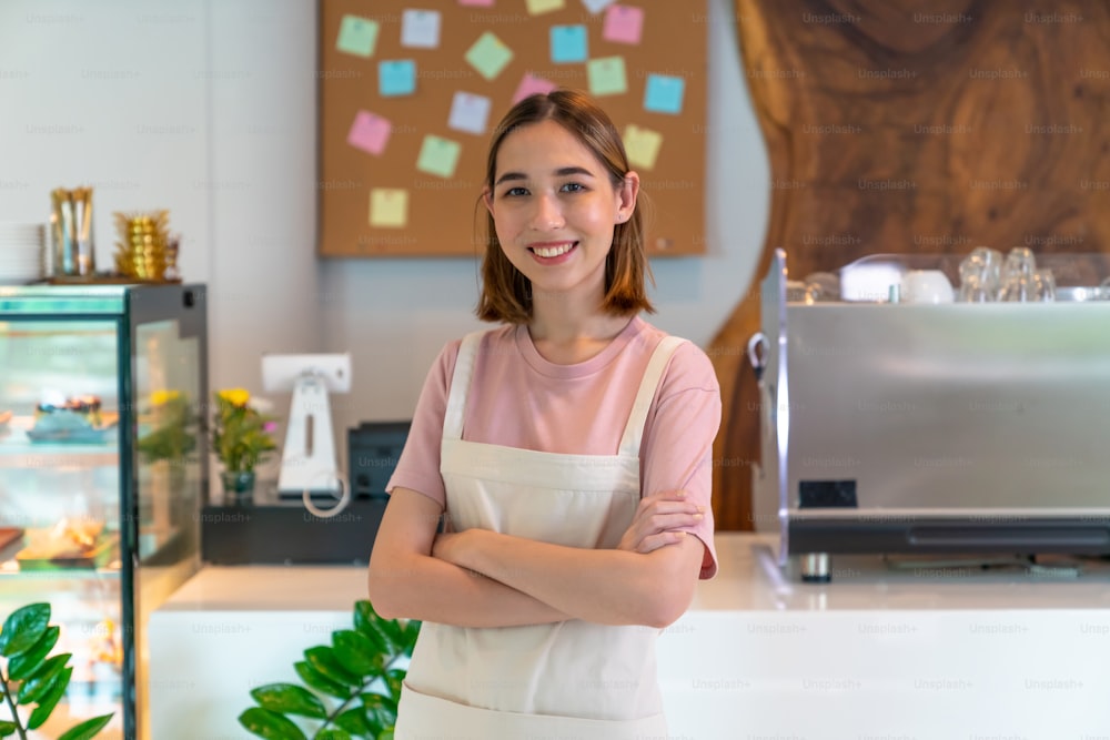 Portrait of Young Asian woman coffee shop waitress standing in front of bar counter crossing arms and look at camera with happiness. Small business restaurant entrepreneur and cafe owner concept