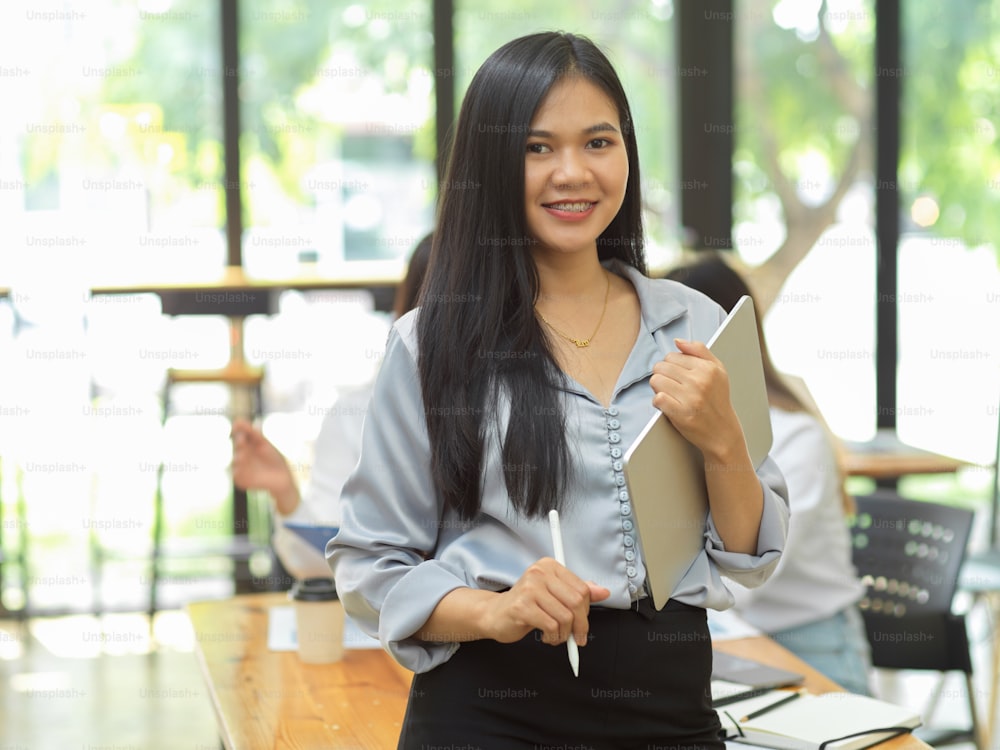 Asian confident businesswoman standing and smiling in front of worktable, holding tablet, meeting room in the background