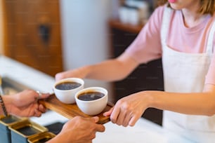 カフェのバーカウンターでホットコーヒーを客に振る舞う若いアジア人女性の喫茶店のウェイトレスのバリスタ。笑顔の女性レストランのレジ係は、クライアントからコーヒーとベーカリーを注文します。中小企業の経営者とパートタイムの仕事のコンセプト