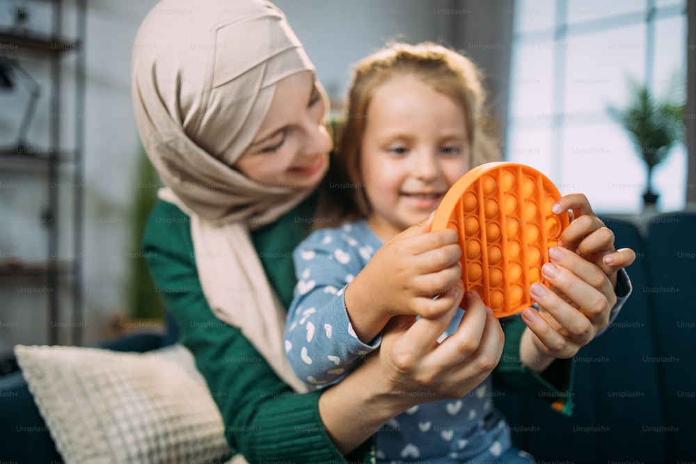 Silicone fidget pop pit close-up. Little happy smiling girl sitting on the sofa at home, playing poppit game together with her pretty Muslim mother. Selective focus on toy.