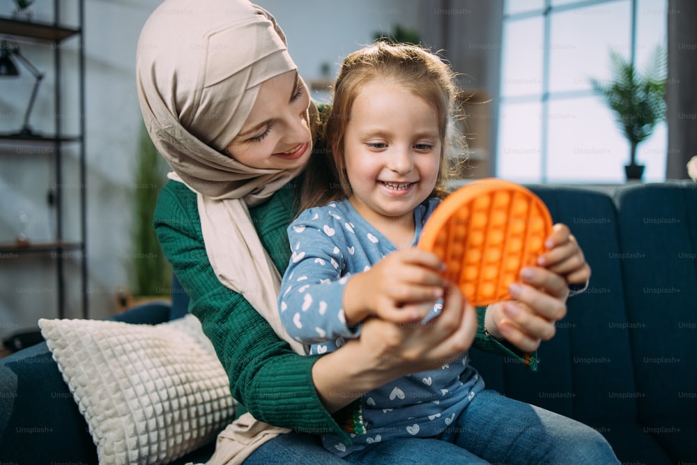 Pädagogische Kinderspiele, Poppit, Antistress-sensorisches Pop-it-Spielzeug. Aufgeregtes, fröhliches, entzückendes kleines Mädchen, das ein beliebtes Anti-Stress-Spiel mit Knöpfen spielt und mit ihrer arabischen Mutter im Hijab auf dem Sofa sitzt