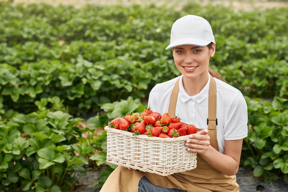 Vista frontale della donna sorridente in maglietta bianca e berretto che tiene una gustosa fragola rossa in un grazioso cesto di vimini su sfondo di piante. Concetto di fragola matura e fresca in serra.
