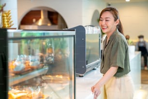 젊은 아름 다운 아시아 여자 커피 숍 직원 바리 스타가 카페에서 일하고 있습니다. 웃는 여성 계산원이 고객으로부터 커피와 빵집을 주문합니다. 중소 기업 소유자 및 아르바이트 개념