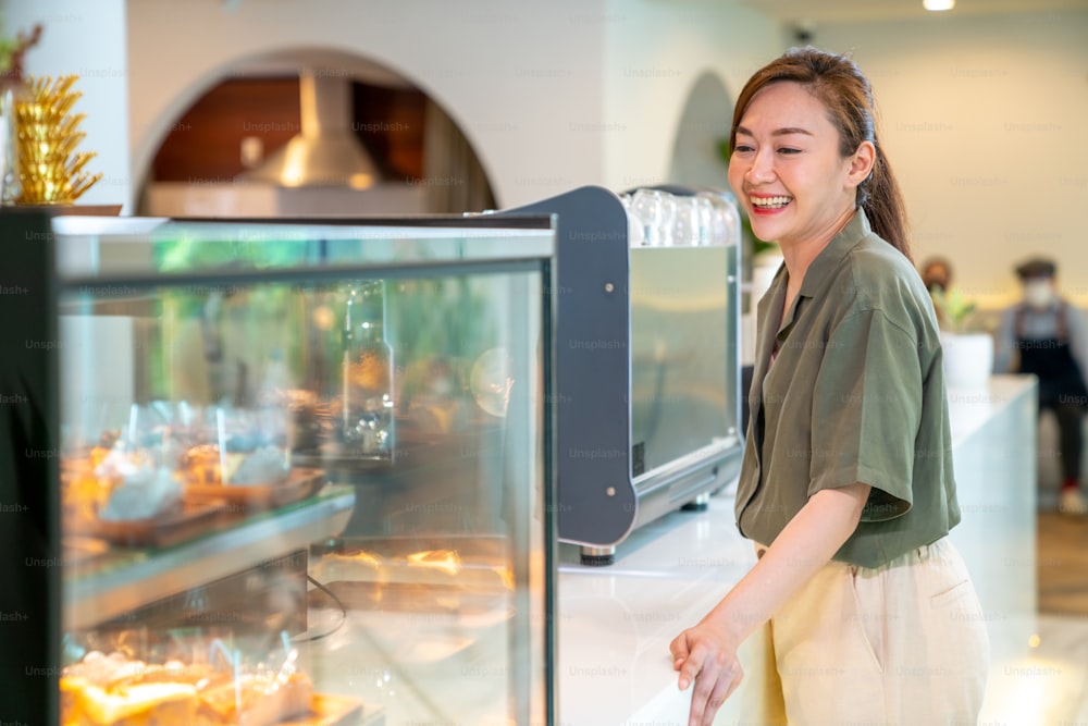 カフェで働く若い美しいアジアの女性コーヒーショップの従業員バリスタ。笑顔の女性レジ係が顧客からコーヒーとベーカリーを注文します。中小企業の経営者とパートタイムの仕事のコンセプト