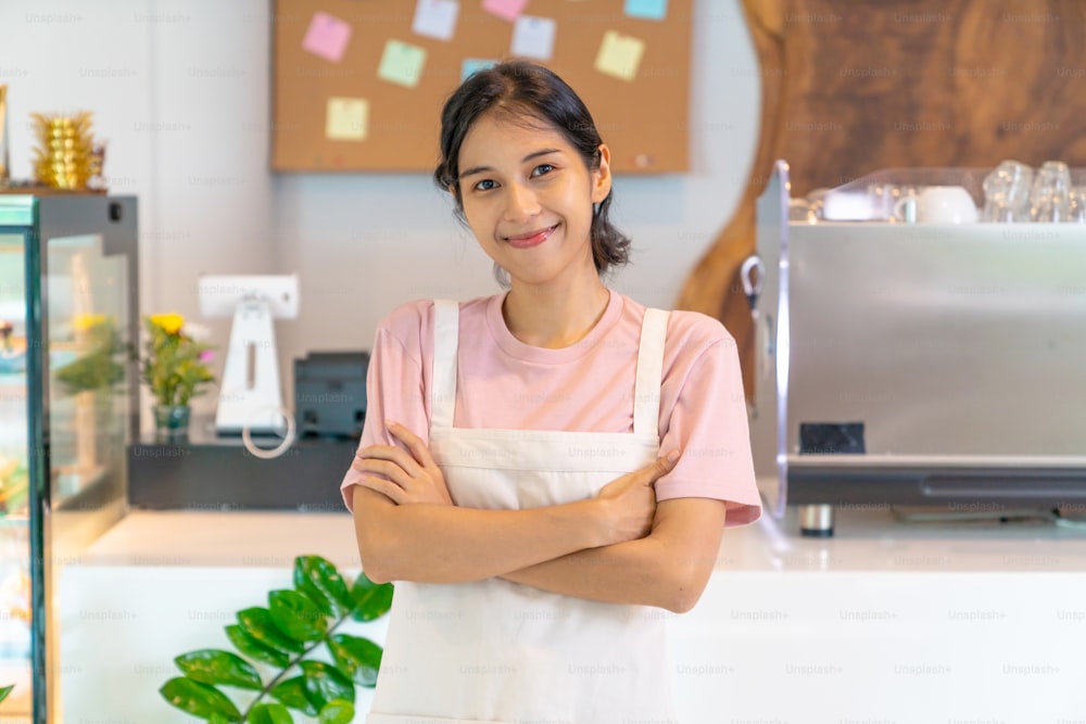 Portrait of Young Asian woman coffee shop waitress standing in front of bar counter crossing arms and look at camera with happiness. Small business restaurant entrepreneur and cafe owner concept