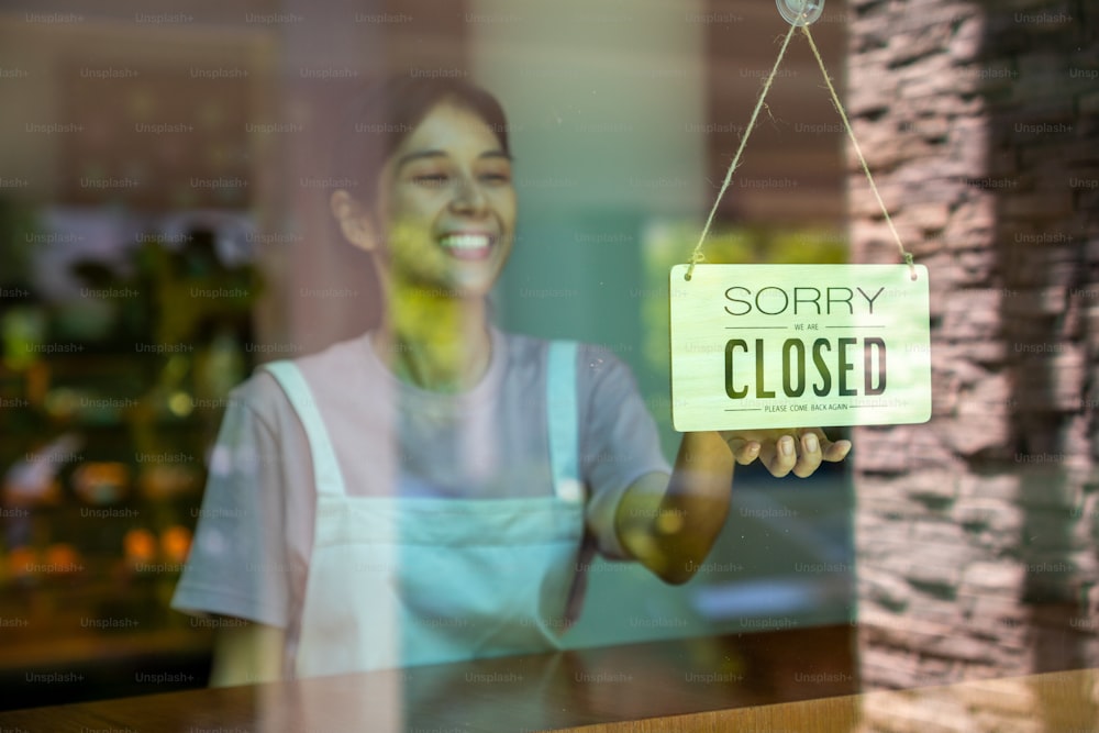 Asian woman coffee shop barista walking to cafe door and turning hanging closed sign. Female waitress preparing restaurant for service to customer. Small business owner and part time job working concept