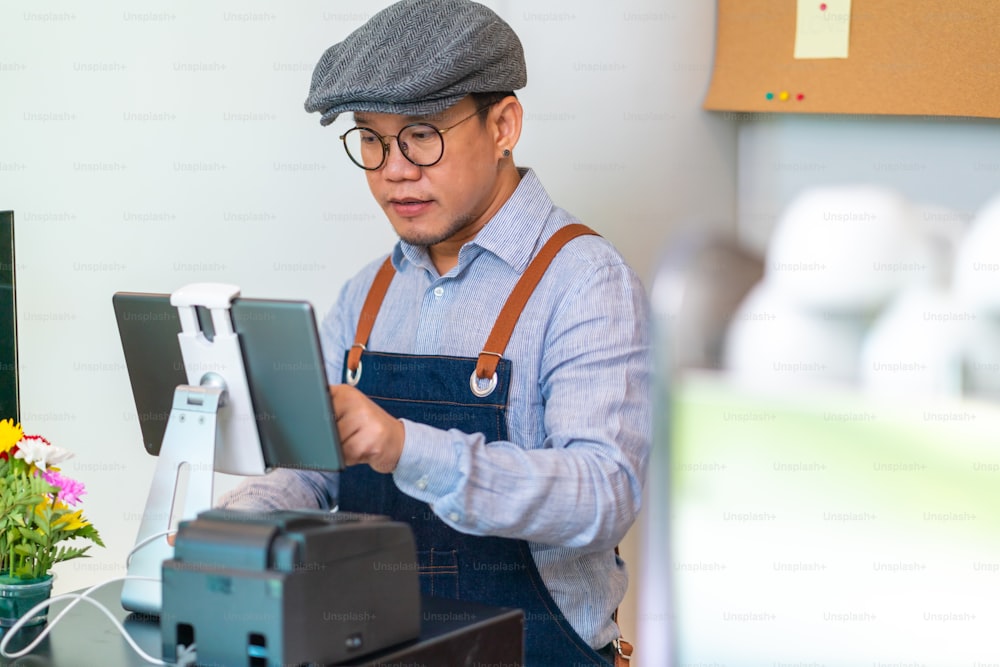 Asian man coffee shop manager walking behind bar counter working on digital tablet checking and counting inventory for ordering product for cafe. Small business restaurant owner entrepreneur concept