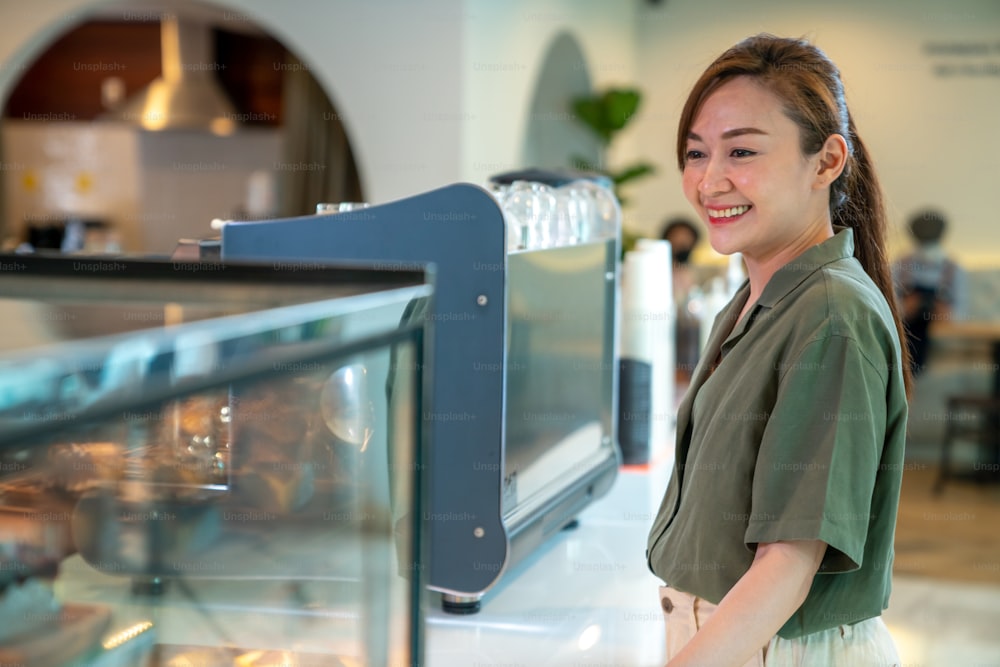 Giovane bella donna asiatica barista dipendente della caffetteria che lavora al bar. Cassiera sorridente che ordina caffè e prodotti da forno dal cliente. Piccolo imprenditore e concetto di lavoro part-time