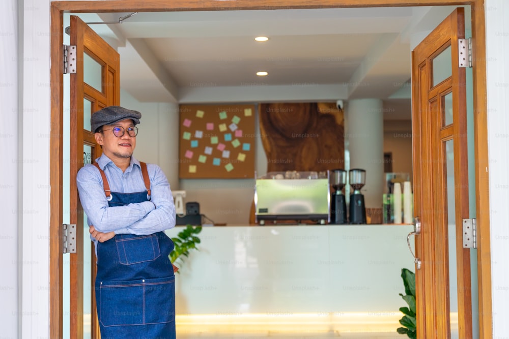 Portrait of Smiling Asian man coffee shop manager open shop door and standing with crossing arms. Male cafe owner waiting for customer with happiness. Small business restaurant entrepreneur concept