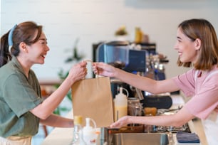 Young Asian woman coffee shop employee barista working at cafe. Smiling female waitress or cashier preparing takeaway order in paper bag to customer. Small business owner and part time job working concept