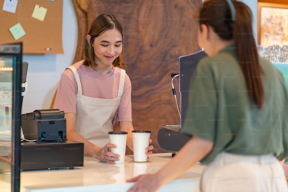 Young beautiful Asian woman coffee shop employee barista working at cafe. Smiling female waitress preparing takeaway order in paper coffee  cup to customer. Small business owner and part time job working concept
