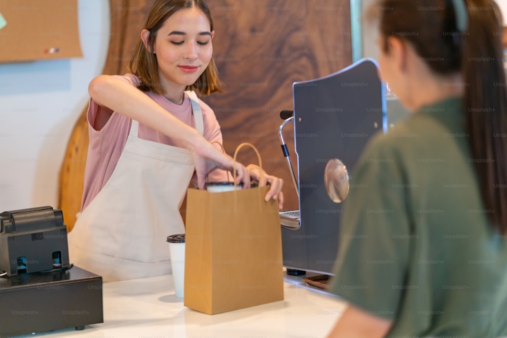 Young Asian woman coffee shop employee barista working at cafe. Smiling female waitress or cashier preparing takeaway order in paper bag to customer. Small business owner and part time job working concept