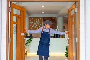 Retrato del gerente de la cafetería del hombre asiático sonriente abre la puerta de la tienda y está de pie con los brazos cruzados. Dueño de una cafetería esperando al cliente con felicidad. Concepto de emprendedor de restaurante de pequeña empresa