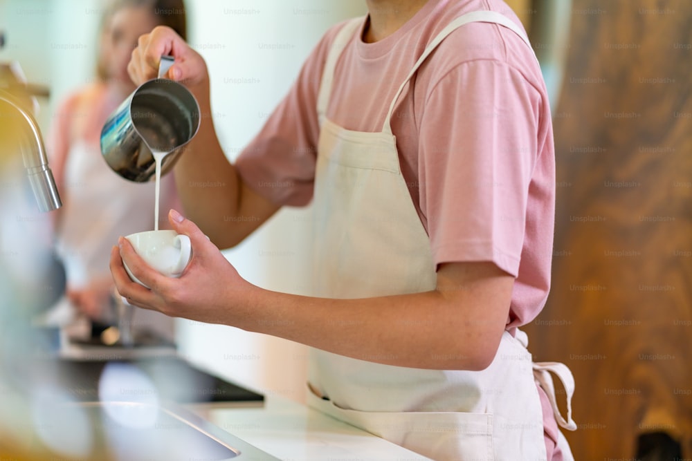 Asiatischer Barista gießt Schaummilch und macht Latte Art Kaffee in Kaffeetasse. Männlicher Coffeeshop-Teilzeitangestellter, der Espresso in der Kaffeemaschine zubereitet. Kleinunternehmer und Teilzeitjob-Arbeitskonzept.