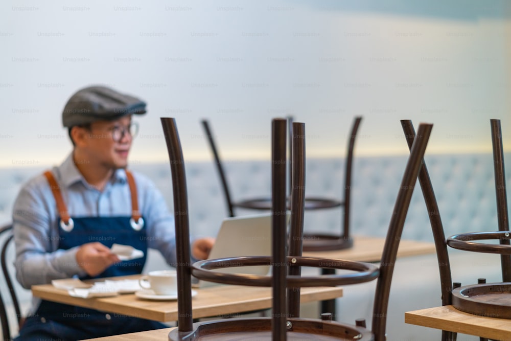 アジア人男性のコーヒーショップのオーナーがテーブルに座り、閉店後にラップトップコンピュータで店の財務請求書を計算しています。ストレスを感じている男性の中小企業のレストランの起業家は、支払いと収入の請求書を数えています。