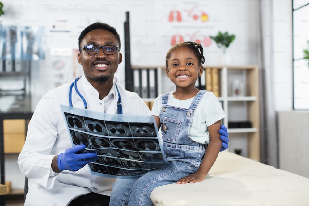 Porträt eines afrikanischen Arztes, der eine kleine Patientin umarmt und den Röntgenscan in der Hand hält. Kinderarzt mit süßem Mädchen, das im Krankenhausbüro sitzt und in die Kamera lächelt.