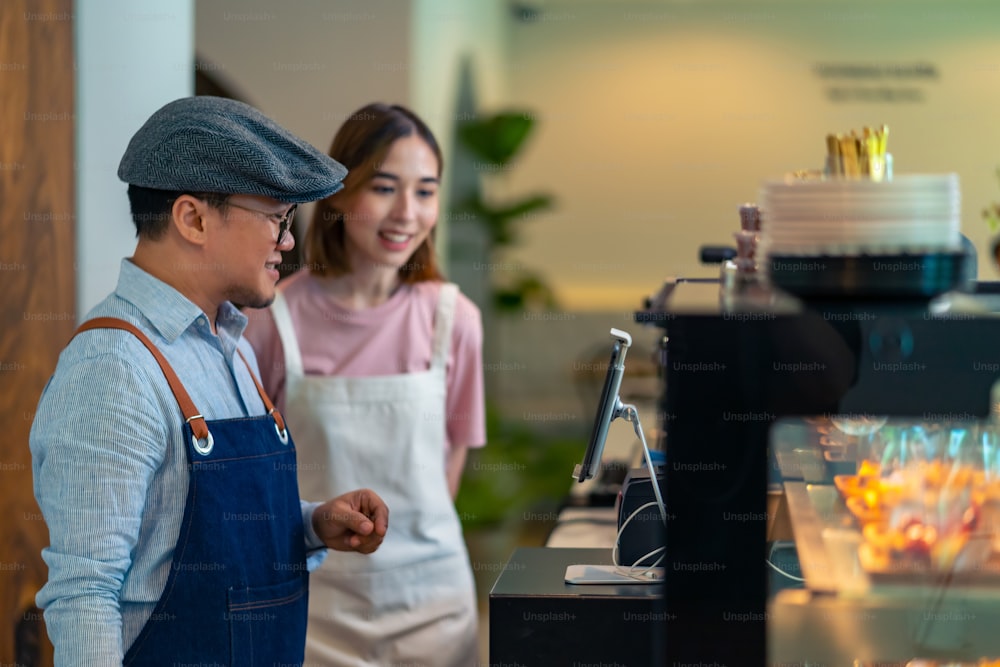 Asian male coffee shop manager teaching young woman staff working on digital tablet. Small business cafe and restaurant owner instruct part time employee preparing service to customers before opening