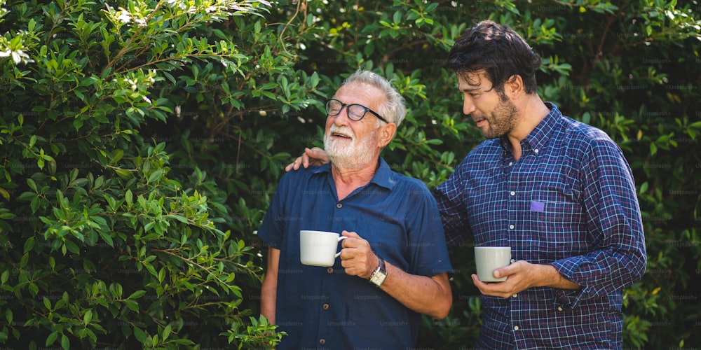 adult son and elderly senior father talking together and holding coffee cup in the garden, happy morning lifestyle of man generation family, love day at home